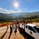 Grupo de pessoas reunidas numa estrada de terra com paisagem montanhosa ao fundo e veículo de apoio branco estacionado.